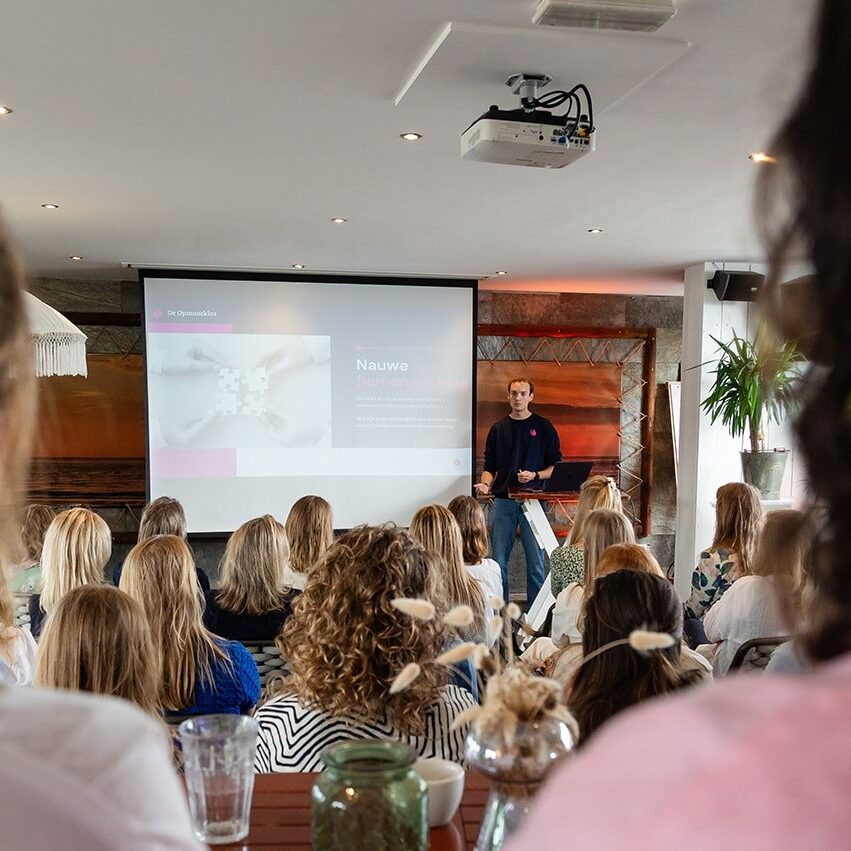 Voorlichting van De Opmaatklas | De Opmaatklas Presenter giving a talk to a seated audience in a conference room with a projector screen behind him